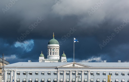 Main green dome of white Helsinki Cathedral and top of Helsinki City Hall with the Flag of Finland in the roof against dark stormy sky.