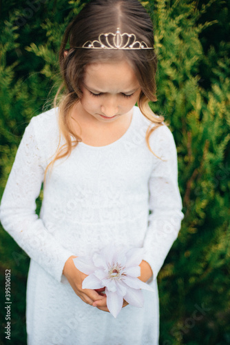 A beautiful little girl in a white dress with a karona on her head Holds a white flower in her hands. Country plants. The child sniffs the scent of flowers. photo