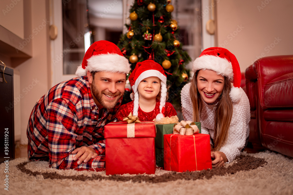 Christmas. Family. Happiness. Portrait of dad, mom and daughter in Santa hats sitting on a couch at home near the Christmas tree, all are smiling. Christmas. Family. Happiness. Portrait of dad, mom and daughter in Santa hats sitting on a couch at home near the Christmas tree, all are smiling.
