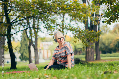 Senior woman resting in the park sitting on the grass, using phone and laptop, and reading a book © Liubomir