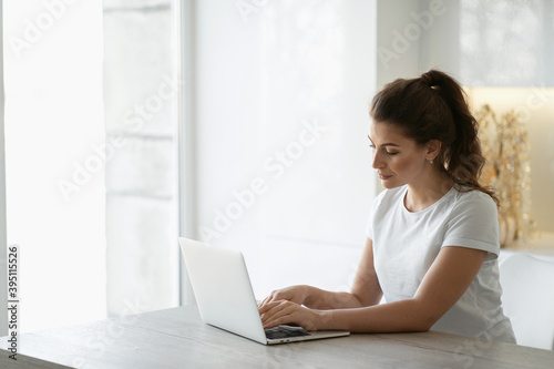 student Manager prints a message and writes a distance learning paper on a computer or laptop. A young woman is sitting at home in the kitchen in front of a gadget, looking at the monitor