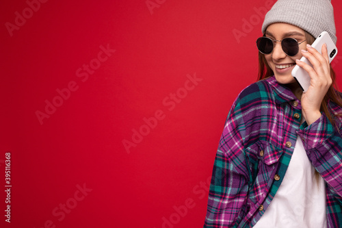 cool girl in a hat and shirt in a cage speaks on the phone. Teenager in hat and sunglasses on red background