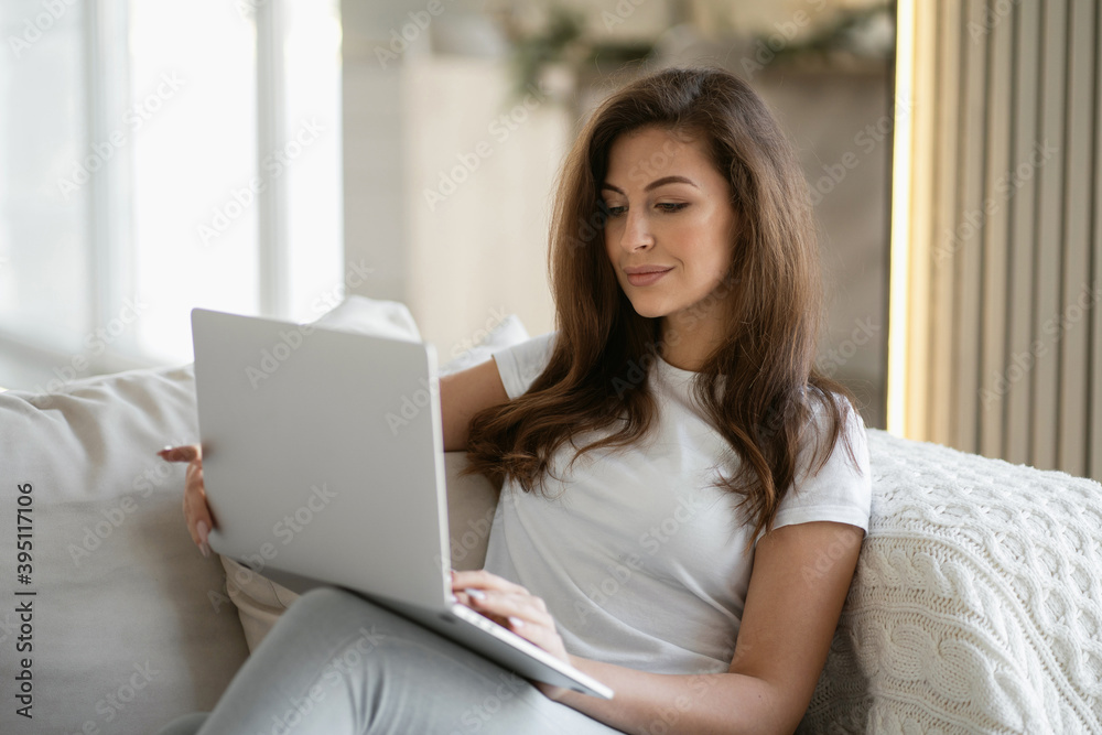 teacher online courses prints a message text to their students, remote work on a laptop computer from your favorite sofa at home. White t-shirt and blue jeans, brunette woman teacher online courses prints a message text to their students, remote work on a laptop computer from your favorite sofa at home. White t-shirt and blue jeans, brunette woman