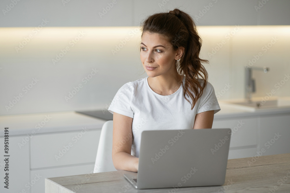 brunette hair in a ponytail beautiful in a comfortable white t-shirt. a woman is typing sitting at home in the kitchen at a laptop computer doing tasks to work online in quarantine. copy space brunette hair in a ponytail beautiful in a comfortable white t-shirt. a woman is typing sitting at home in the kitchen at a laptop computer doing tasks to work online in quarantine. copy space