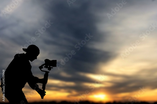 Silhouette of videographer is filming with cinema gimbal video dslr at sunset , professional video, videographer in events. Cinema lens on gimbal. Medium shot from right side. Film or cameraman school