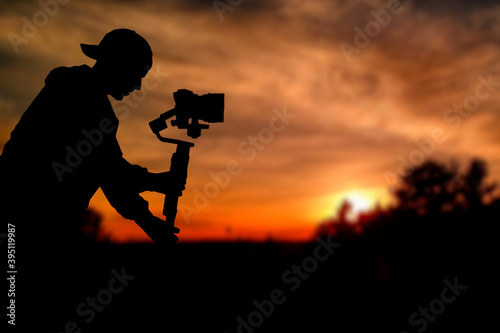 Silhouette of videographer is filming with cinema gimbal video dslr at sunset , professional video, videographer in events. Cinema lens on gimbal. Medium shot from right side. Film or cameraman school