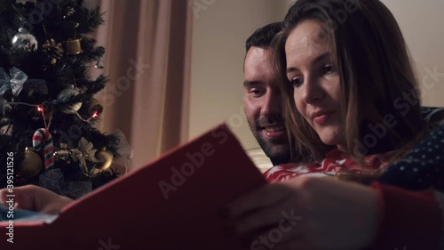 Beautiful couple sitting on the sofa at Christmas looking at a photo album. Large green Christmas tree with toys and garland.