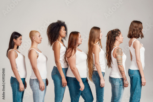 Group of beautiful diverse young women wearing white shirt and denim jeans looking aside while posing, standing isolated over grey background