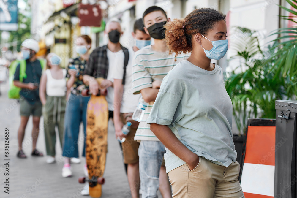 Young african american woman wearing mask waiting, standing in line with other people, respecting social distancing to collect takeaway order from the pickup point during lockdown