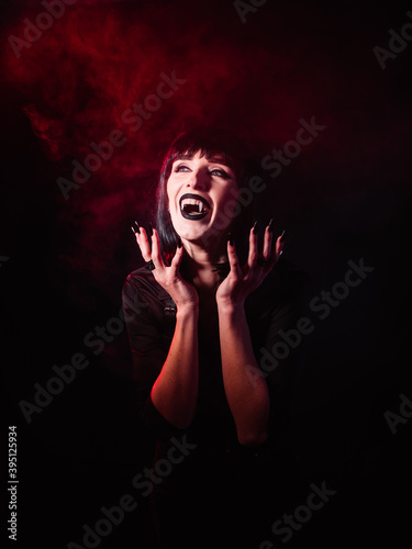 Girl on a black background with red light and artificial smoke in the image of a vampire. She laughs with her mouth open, showing her fangs.