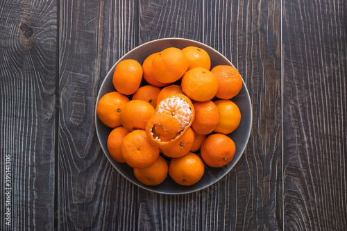a plate of ripe orange tangerines on a wooden table, one peeled tangerine on top
