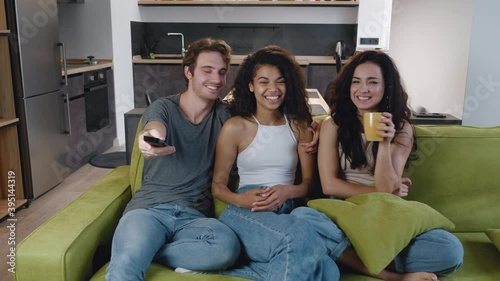 Group of three friends talking, laughing and watching TV together while sitting on the couch. Mixed race young people have a party at home