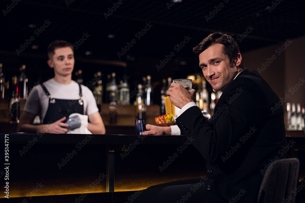 a handsome man poses at the bar with a cocktail in his hands against the background of the bartender and bottles.