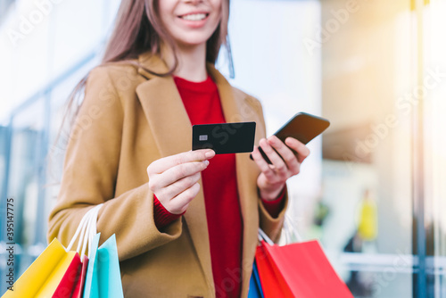 Cropped shot of elegant young woman using smartphone to check a balance on her credit card while standing with a bunch of colorful shopping bags in hands at the mall
