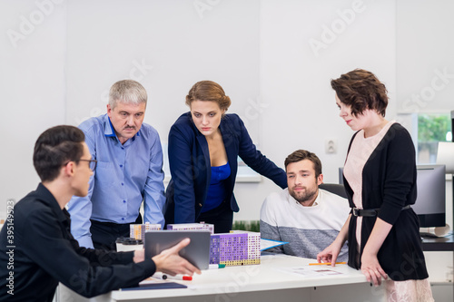 Office business meeting. The team is sitting at a table in a luminous white open space. The senior grey haired boss is up and listening carefuly to his younger employee, who is showing his photo
