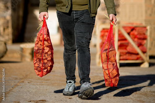 A man carries two red net sacks containing firewood.