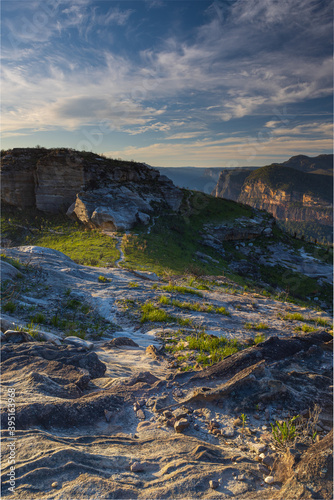 Trail in mountains leads along to the cliff. Mountain walls in the background highlighted by the setting sun under blue evening sky