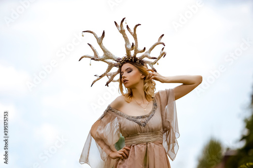 Portrait of Young Beautiful Caucasian Girl Posing With Lifted hand in Light Dress With Decorative Deer Horns Outdoors Against Bright Skies.