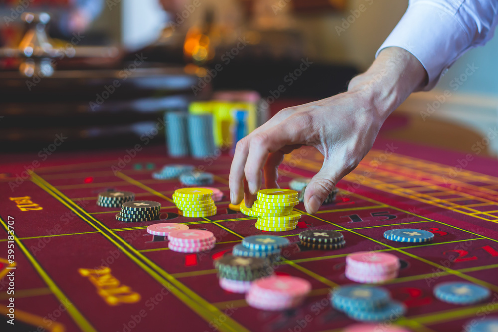 A close-up vibrant image of multicolored casino table with roulette in motion, with casino chips. the hand of croupier, mone and a group of gambling rich wealthy people in the background A close-up vibrant image of multicolored casino table with roulette in motion, with casino chips. the hand of croupier, mone and a group of gambling rich wealthy people in the background