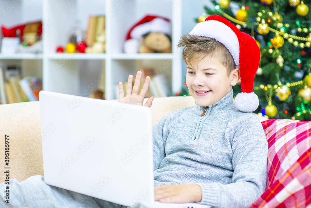 A boy  sits at home on the sofa against the background of a Christmas tree in a Santa hat and makes a video call to his family to wish them Christmas.