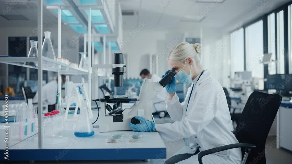 Modern Medical Research Laboratory: Portrait of Female Scientist Wearing Face Mask Looking Under Microscope, Analysing Test Samples. Advanced Scientific Lab for Medicine, Microbiology Development Modern Medical Research Laboratory: Portrait of Female Scientist Wearing Face Mask Looking Under Microscope, Analysing Test Samples. Advanced Scientific Lab for Medicine, Microbiology Development