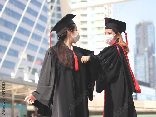graduated women in graduation gowns and medical face masks, elbows greeting to avoid the spread of virus.. Graduation Class of 2020. Covid-19 prevention and new normal concept.