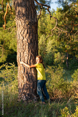 A girl with long hair and a yellow t-shirt hugs a large pine trunk in summer. The concept of unity with nature