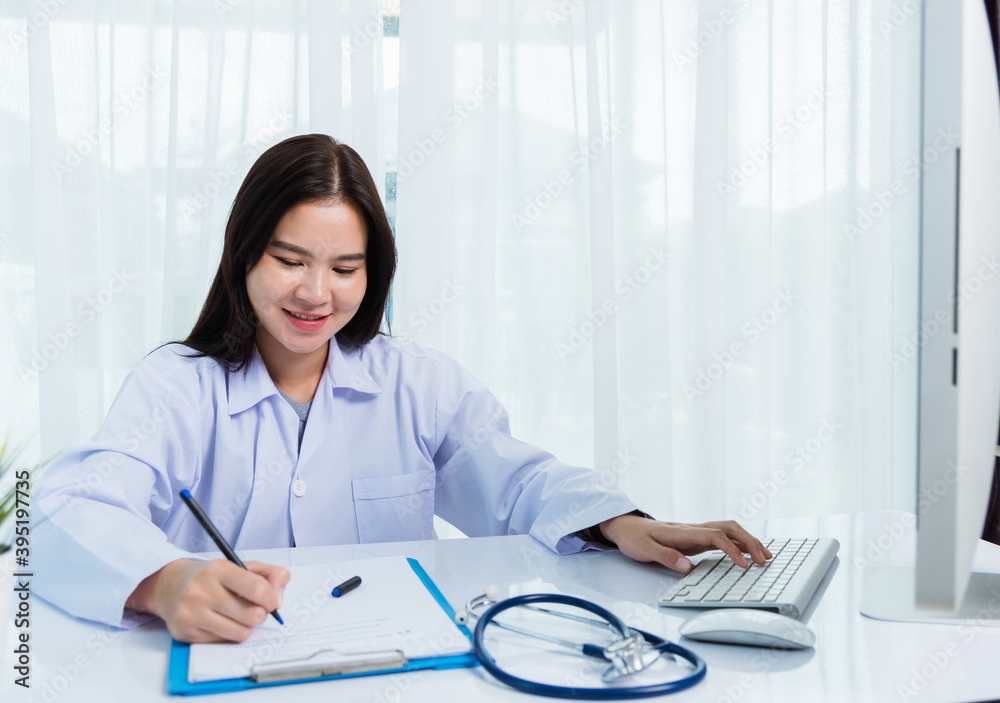 Asian doctor young beautiful woman smiling using working with PC desktop computer and her writing something on paperwork or clipboard white paper at hospital desk office, Healthcare medical concept Asian doctor young beautiful woman smiling using working with PC desktop computer and her writing something on paperwork or clipboard white paper at hospital desk office, Healthcare medical concept