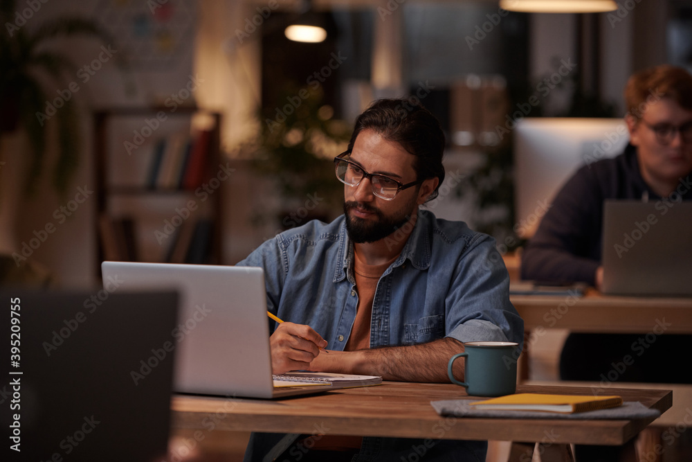 Young bearded businessman in eyeglasses making notes and working on laptop at the table at office
