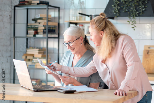 Senior woman using laptop for websurfing in her kitchen. middle-aged daughter helps her mother with documents. Mature lady sitting at work typing a notebook computer in an home office. photo