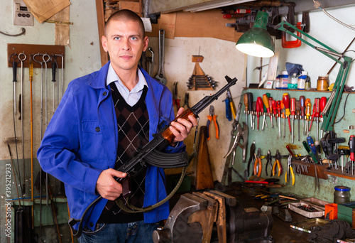 Portrait of confident master of weapons repair workshop standing near workplace with Kalashnikov assault rifle in hands