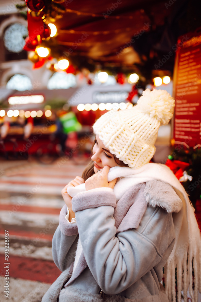 Happy woman enjoys winter holidays. Festive Christmas market on the background. Model wearing stylish winter jacket, knitted beanie hat, scarf.
