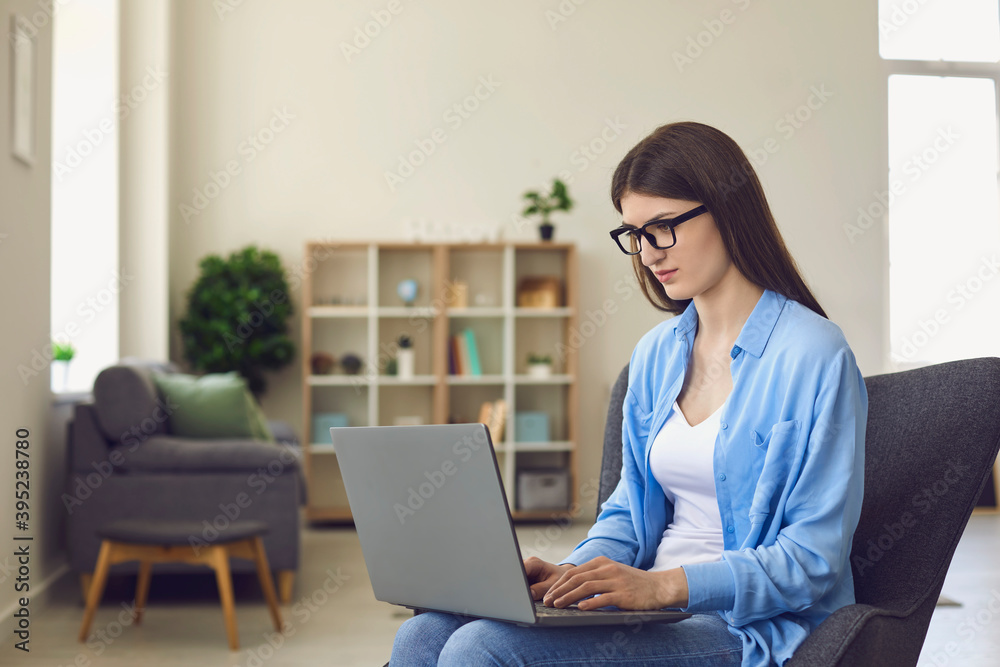 A girl in glasses works with a laptop online looks at the camera while sitting in a chair in the living room. A girl in glasses works with a laptop online looks at the camera while sitting in a chair in the living room.