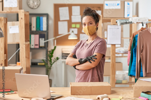 Portrait of young woman in protective mask looking at camera while packing parcel before delivery