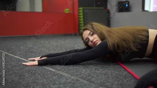 A young fit athletic girl with loose hair in a black top, black leggings does body stretching exercises on the mat in the fitness room. Healthy lifestyle. stretching.