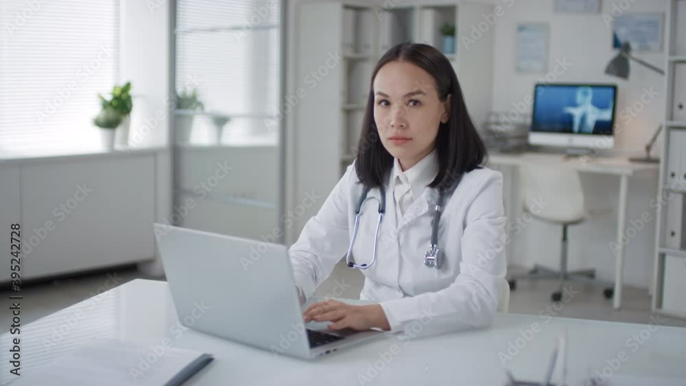 Portrait of modern Asian female medical specialist sitting at desk in office room working on laptop then looking at camera
