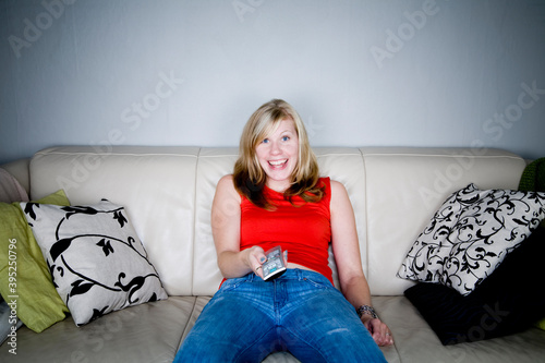 Happy blonde woman in red top and jeans with remote control sitting alone in beige couch.