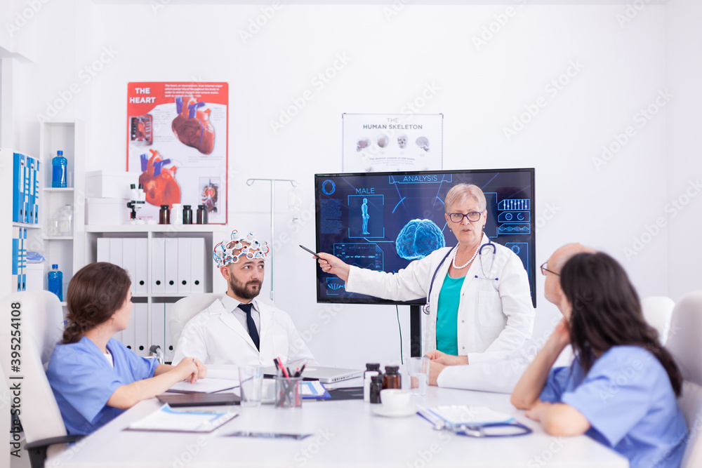 Scientist doing brain analysis using headset with sensors on doctor in hospital meeting room. Monitor shows modern brain study while team of scientist adjusts the device.