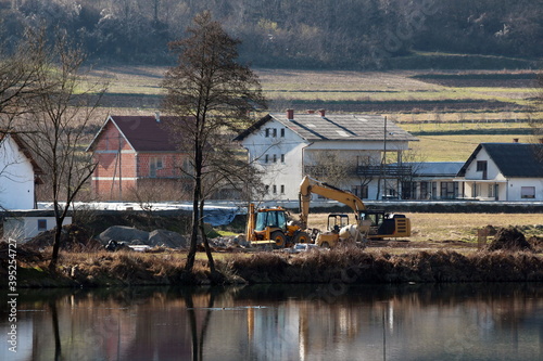 Industrial backhoe loader next to compact excavator and small industrial mixer at local levee construction site next to calm river surrounded with construction material and suburban family houses