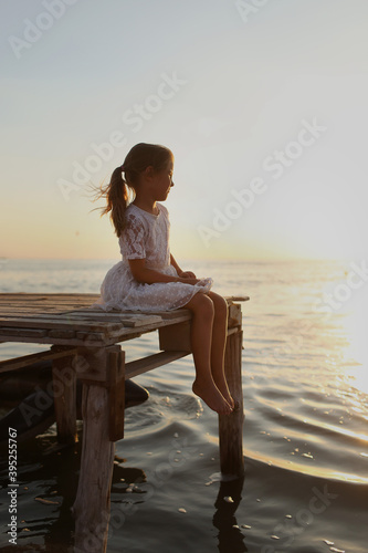 A child sits on the bridge in a light summer dress and looks at the sunset at the sea on the horizon