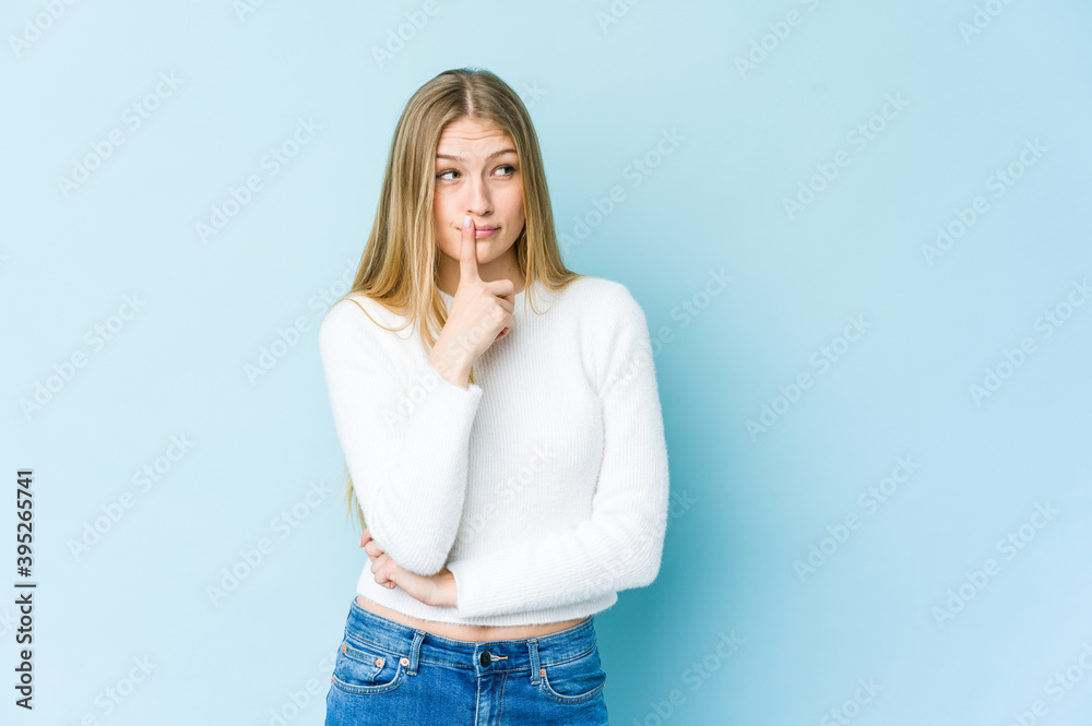 Young blonde woman isolated on blue background looking sideways with doubtful and skeptical expression.