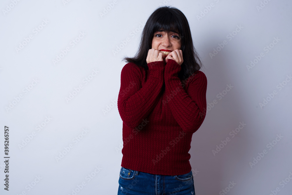 Anxiety - Young beautiful Caucasian woman wearing red T-shirt against white wall, covering his mouth with hands scared from something or someone bitting nails.