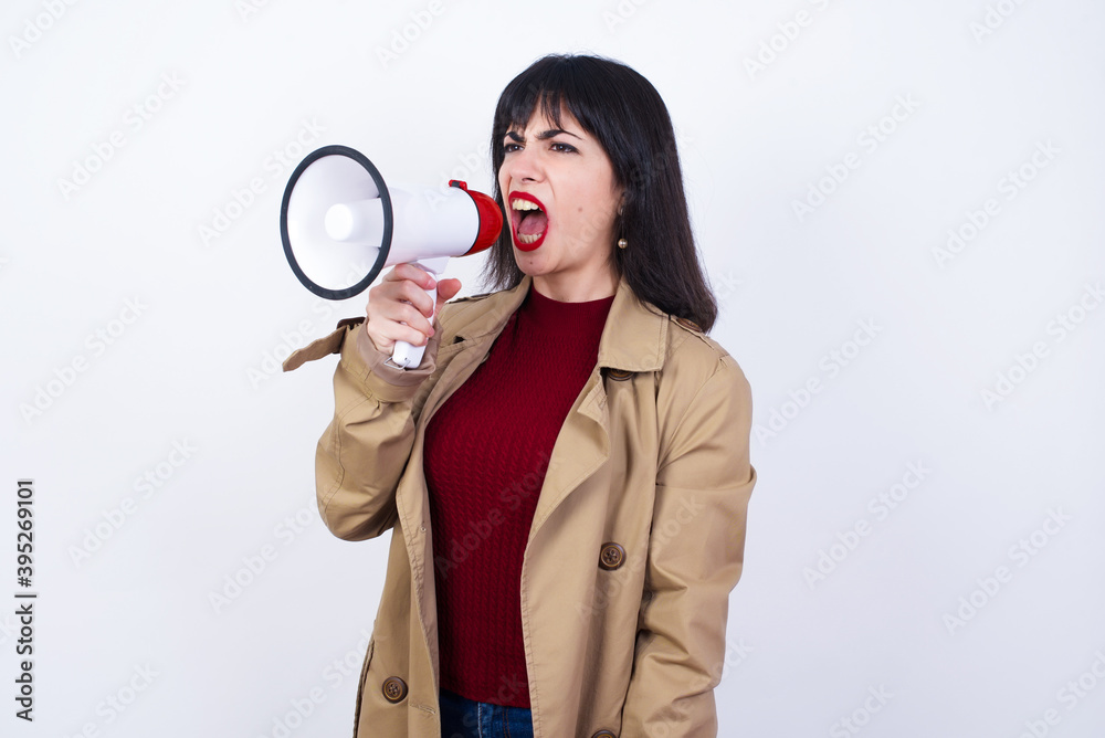 Young beautiful caucasian businesswoman standing against white background, communicates shouting at smartphone with megaphone. Marketing and sales concept. Young beautiful caucasian businesswoman standing against white background, communicates shouting at smartphone with megaphone. Marketing and sales concept.