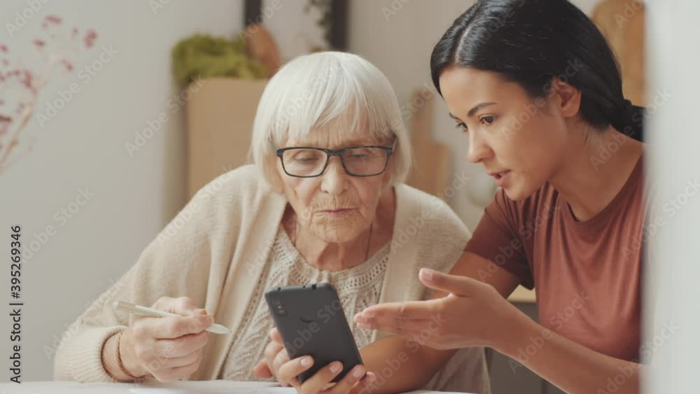 Young Asian female volunteer explaining how to use modern smartphone to senior Caucasian woman and answering her questions while sitting together at home Young Asian female volunteer explaining how to use modern smartphone to senior Caucasian woman and answering her questions while sitting together at home