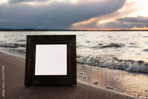 Close-up horizontal photo of a single dark brown wooden squre empty frame for photo on a sea shore with cloudy sky on the background