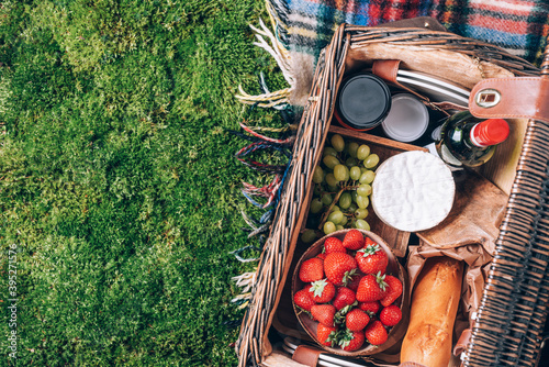 Picnic set with fruit, cheese, honey, strawberries, grapes, baguette, wine, wicker basket for picnic on plaid over green grass. Top view. Copy space. Summer picnic time, family lunch. Romantic picnic