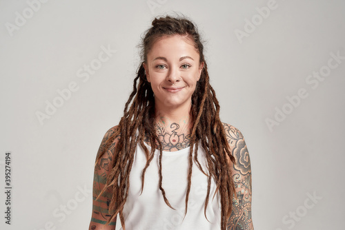 Portrait of young tattooed caucasian woman with dreadlocks wearing white shirt, smiling at camera while posing isolated over grey background