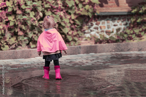 A nice little girl in a rose raincoat and pink rubber boots jumps on puddles with splashes and rejoices. Park, nature, outdoors. Universal Children's Day. childhood