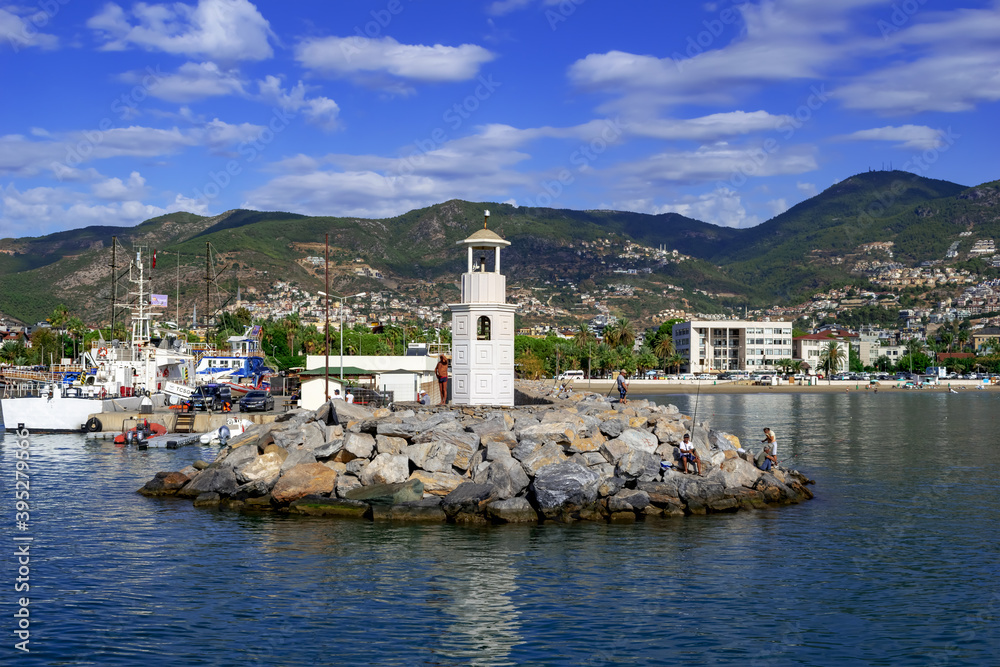 Turkey, Alanya - October 22, 2020: Men fish on rocks near a small lighthouse in the port of Alanya. Cityscape of turkish town with beach, ships and mountains on the horizon