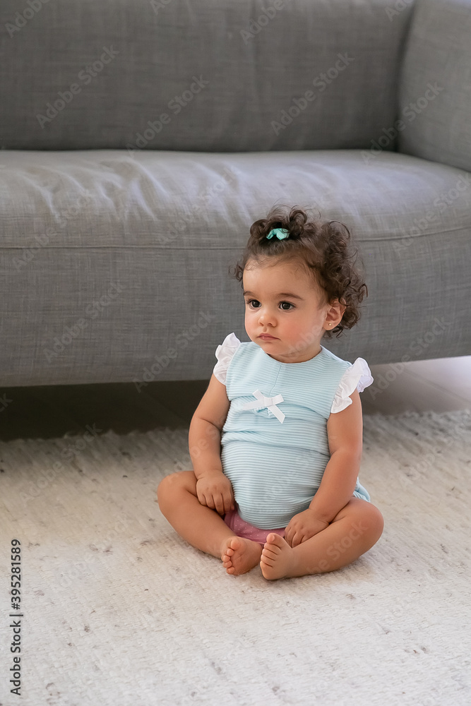 Sweet dark curly haired baby girl wearing pale blue cloth, sitting on floor at home, looking away. Vertical shot. Kid at home and childhood concept Sweet dark curly haired baby girl wearing pale blue cloth, sitting on floor at home, looking away. Vertical shot. Kid at home and childhood concept